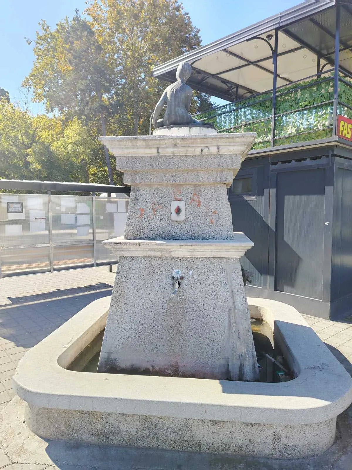 Znak karo na česmi i ženska figura gledana s leđa, uz autobusko stajalište